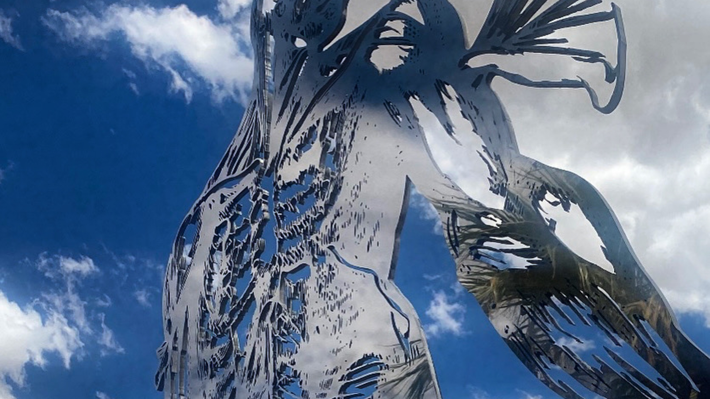 Cor Fabrica, mirror-polished stainless steel sculpture by C Fodoreanu, photographed from below against blue sky and scattered cumulus; the figure's torso and outstretched arm with wing-like ginkgo-leaf cuts read as silver-edged anatomical line-drawing dissolving into the sky.