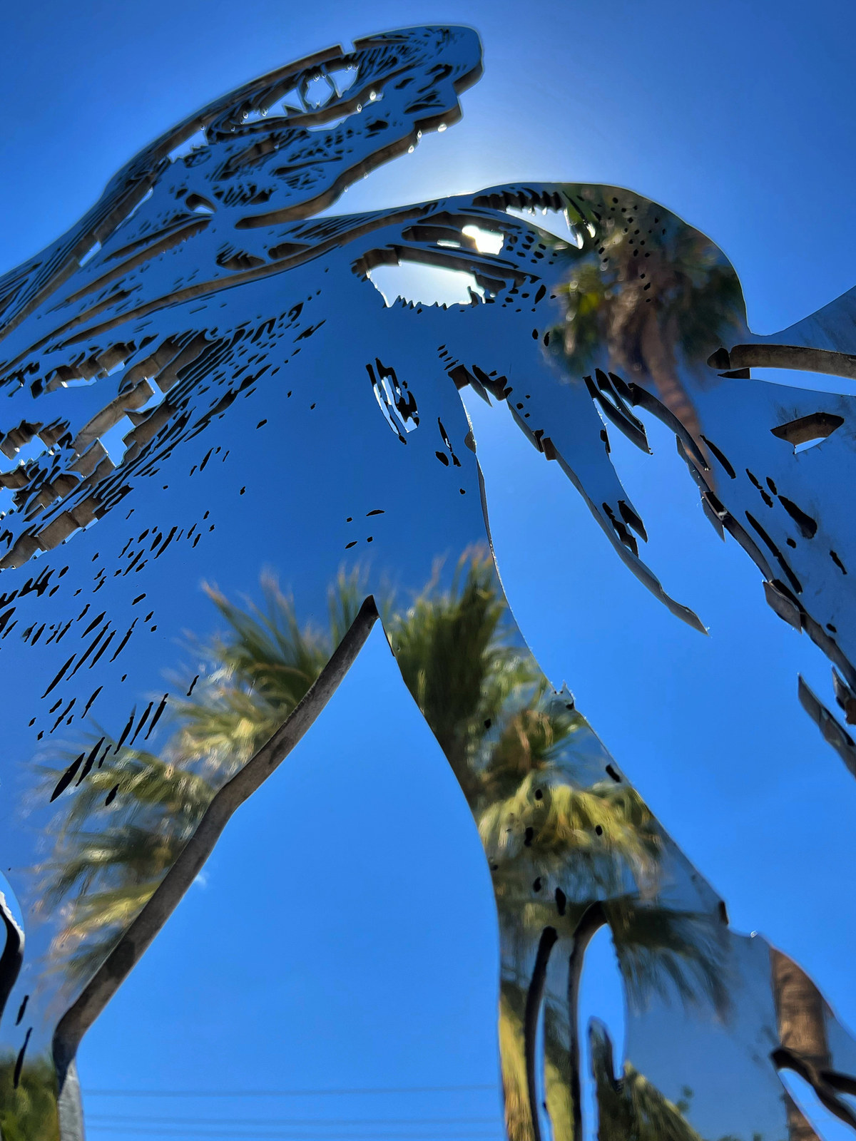 Cor Fabrica, installation view at Chocolate Drop, Twentynine Palms, 2023 — looking up the length of the figure against deep blue sky, palms reflected in the polished steel