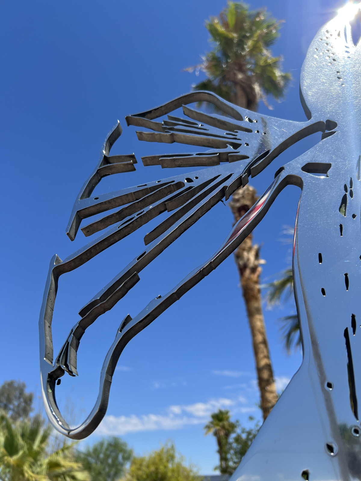 Cor Fabrica, installation view at Chocolate Drop, Twentynine Palms, 2023 — detail of the polished steel wing and ribcage against blue sky with palms