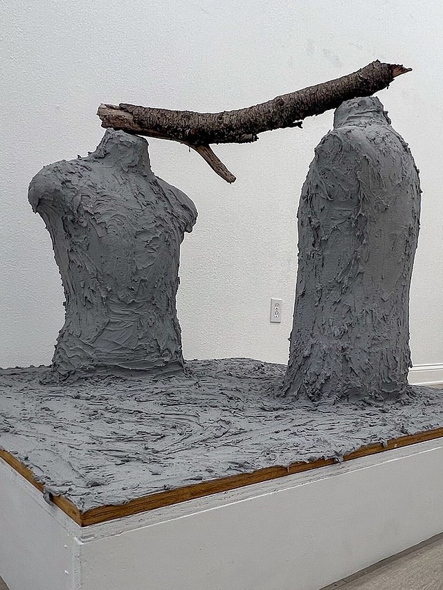 lorem ipsum — two hand-applied cement torsos joined at the shoulders by a barked tree branch, set on a cement-covered pedestal in a gallery, LOS/NR 2025.
