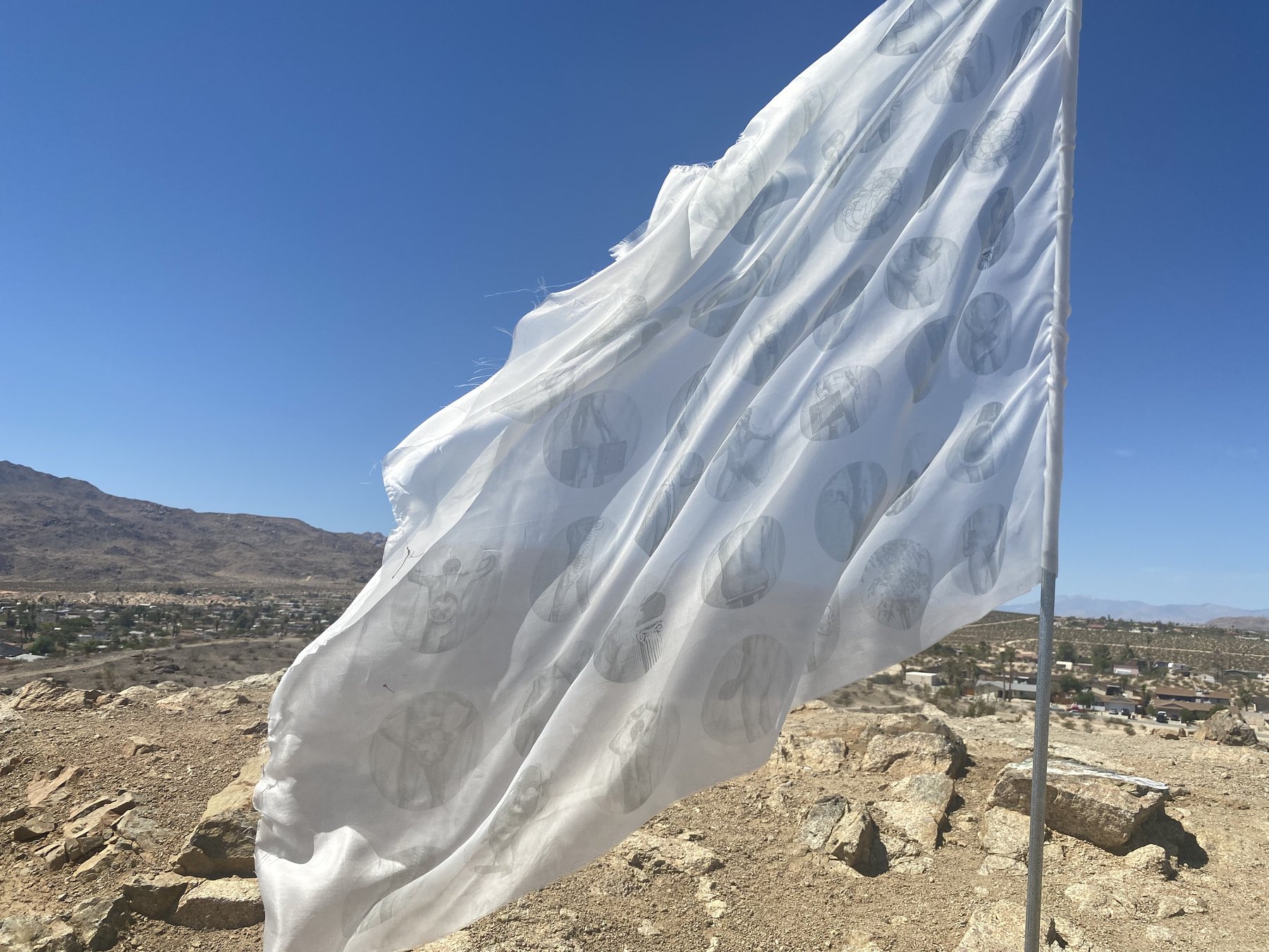 the white flag installed at Chocolate Drop, Twentynine Palms, CA, 2021 — silk flag flying from metal pole in desert wind, the printed body figures visible through the translucent fabric