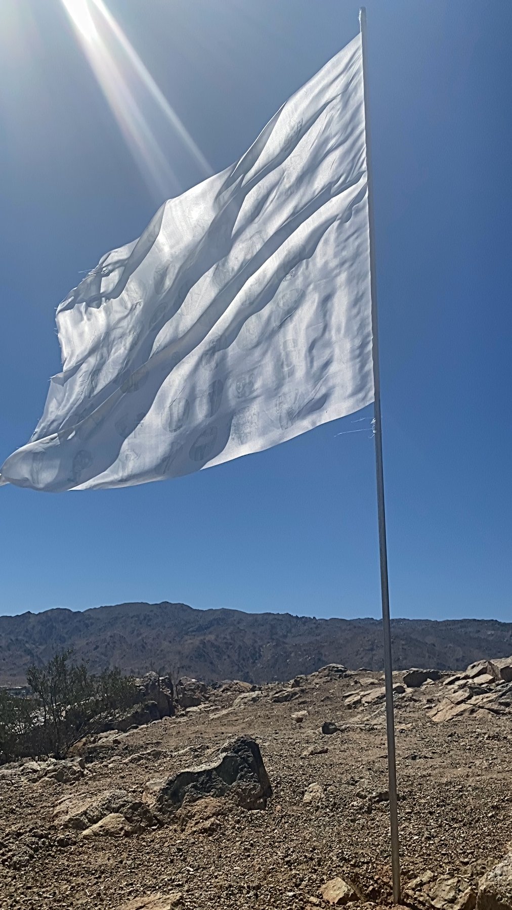 the white flag installed at Chocolate Drop, Twentynine Palms, CA, 2021 — flag on pole against sunlit blue sky and distant mountains