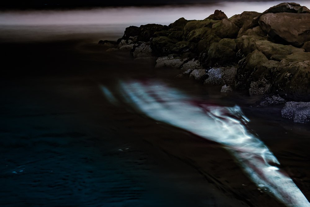 night swim #10, luminous form rippling on the water beside dark coastal rocks, 2020