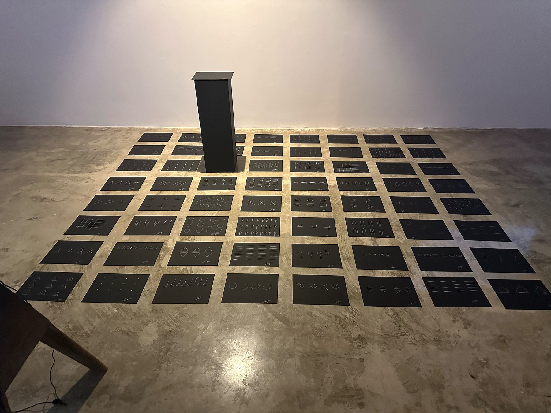 the giveaway, installation view at Ode to the Lake Sacalaia, Ronald H. Silverman Fine Arts Gallery, Cal State LA, 2023 — full floor grid of black panels with white chalk glyphs lit by overhead light, a black pedestal rising vertically from the middle of the grid
